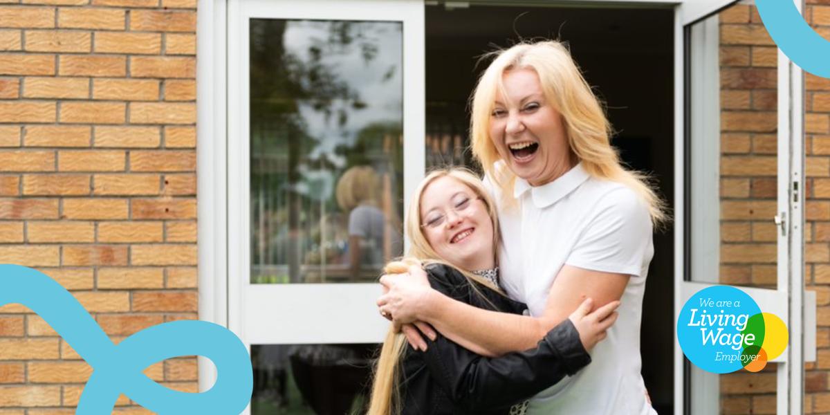 Two people smile at the camera, hugging each other. They are outside with a building behind them.