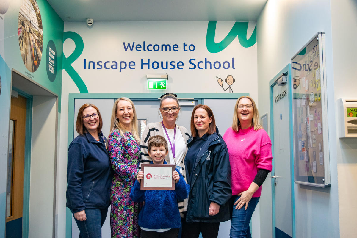 The Nurture team and a pupil, holding the Award certificate and posing for a photo in front of the "Welcome to Inscape House School" wall