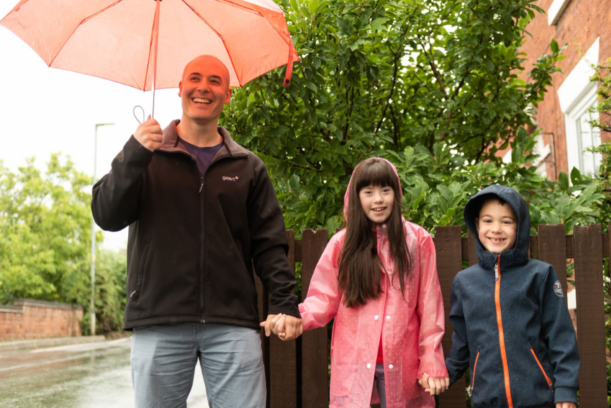 a man with two children smiling at the camera. They are holding hands and the man is holding an umbrella.
