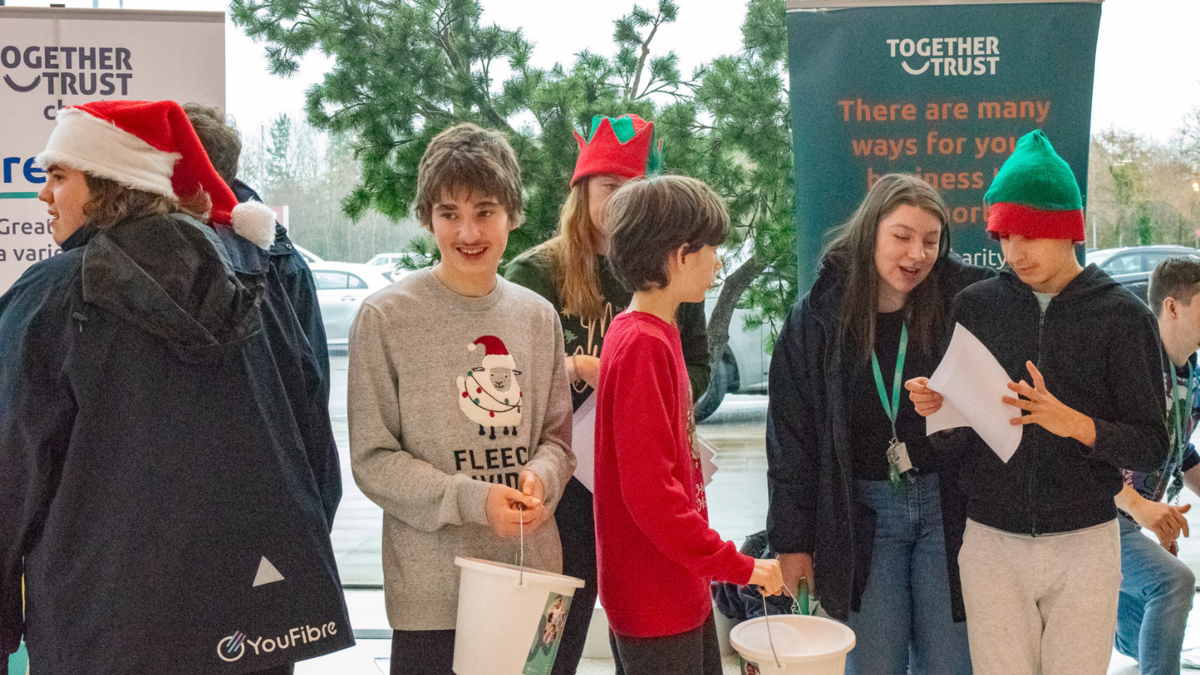 A group of students wearing christmas hats stood inside a shopping centre. They are holding music sheets and singing. 