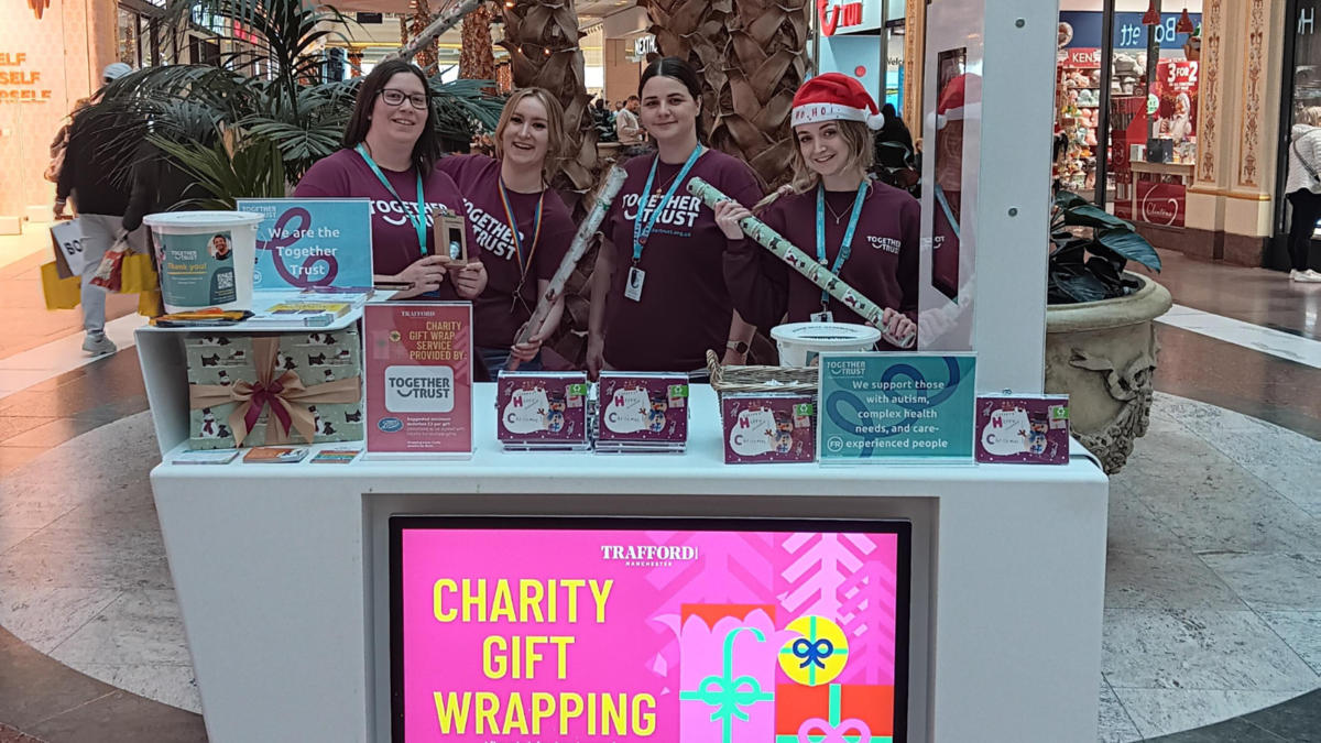 A group of four women stood behind a counter that reads "Charity gift wrapping". They're holding rolls of wrapping paper smiling. 