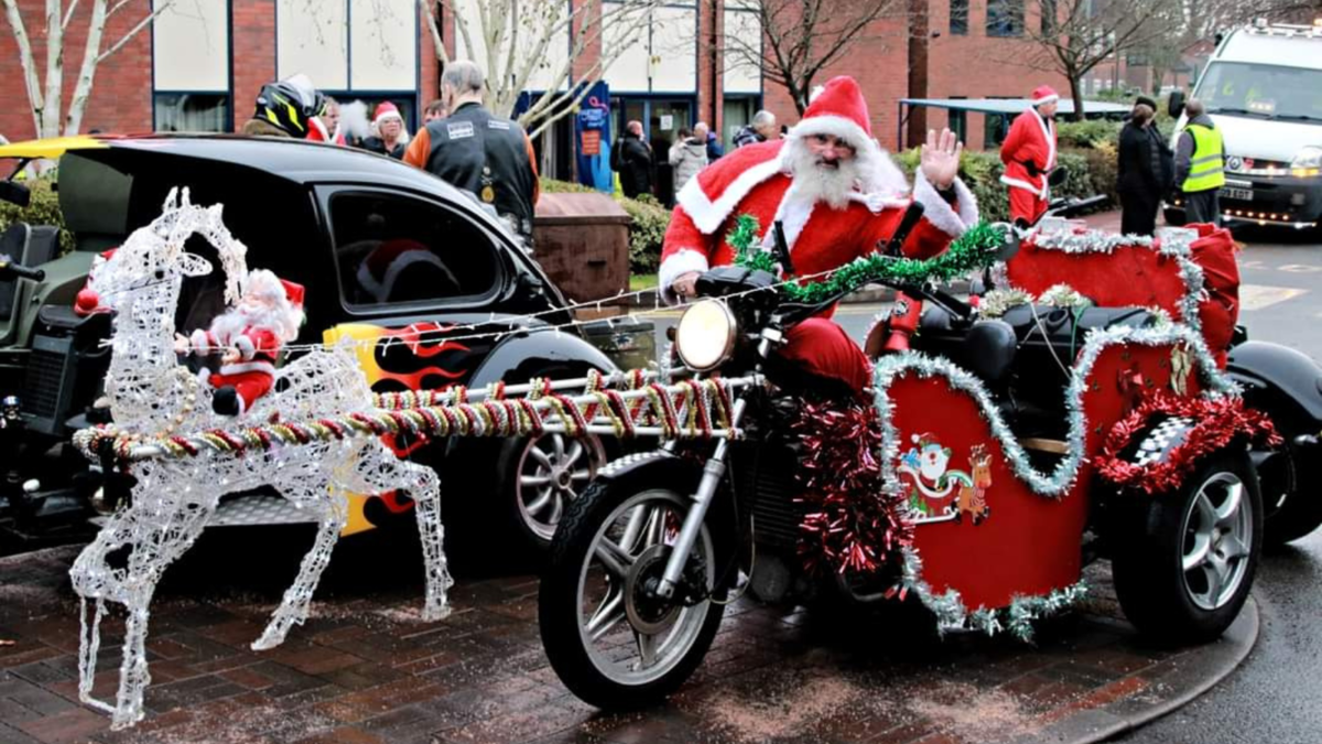 Santa Claus next to a motorbike decorated to look like a sleigh being pulled by a lit up reindeer. 