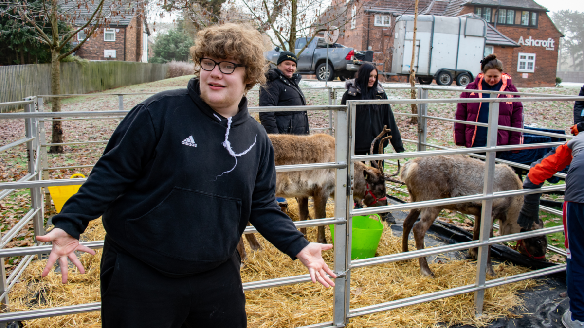 Teenage boy standing in front of the reindeer enclosure pointing at the 2 reindeer. 