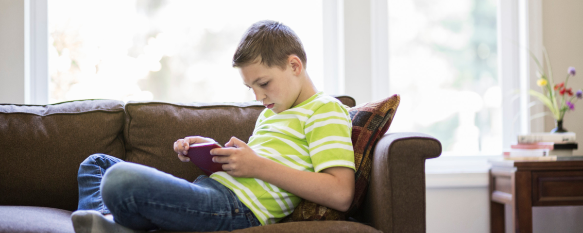 Boy sat crosslegged on a sofa, looking down at a game.