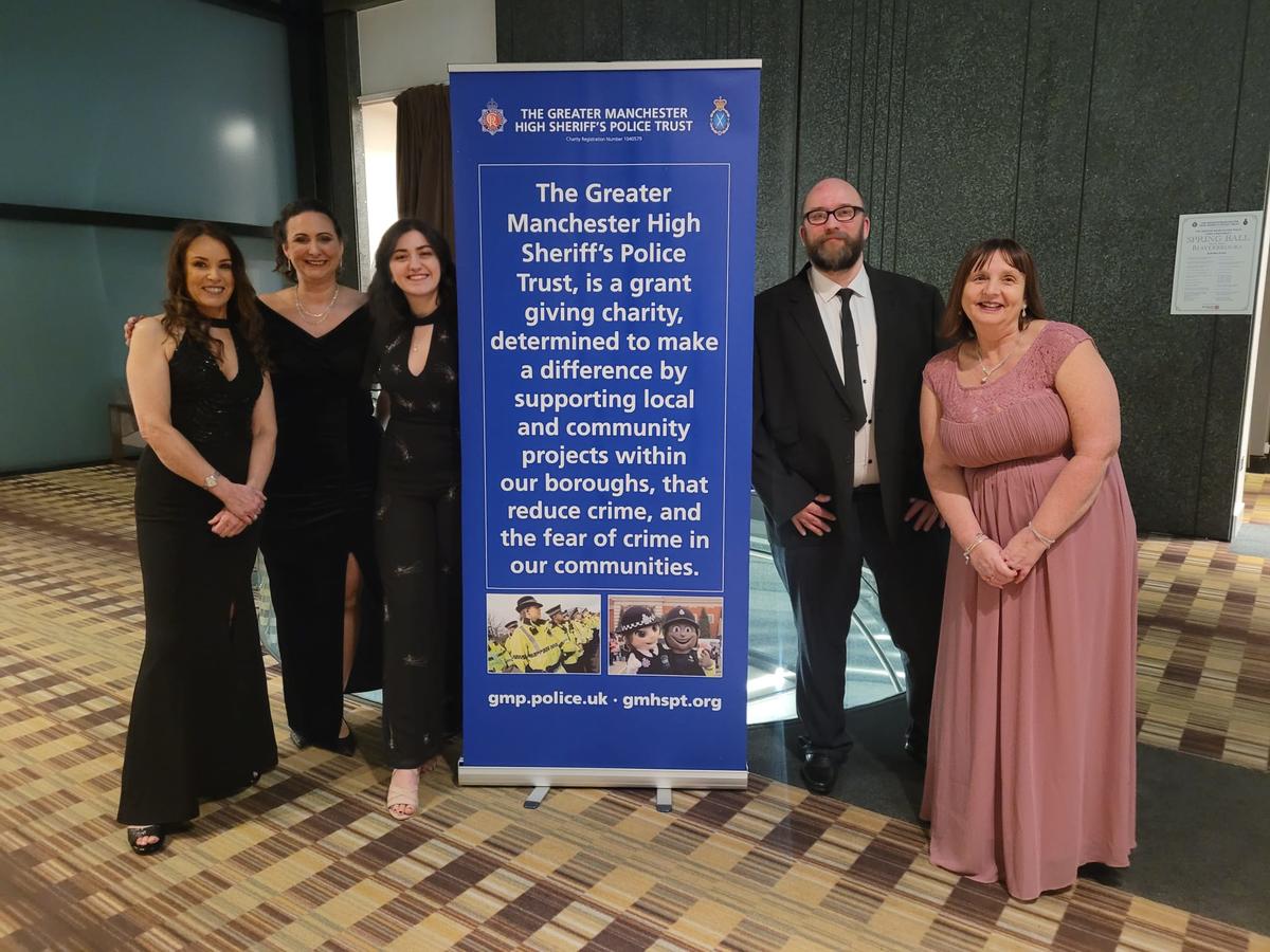 5 members of staff dressed in elegant clothes standing next to a banner that reads "The greater manchester high sheriff's police trust is a grant giving charity determined to make a difference by supporting local and community projects within our boroughs, that reduce crime and the fear of crime in our communities" 