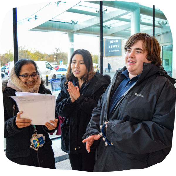 A student and two teachers singing inside John Lewis. They are holding a sheet of lyrics for the songs, and smiling.