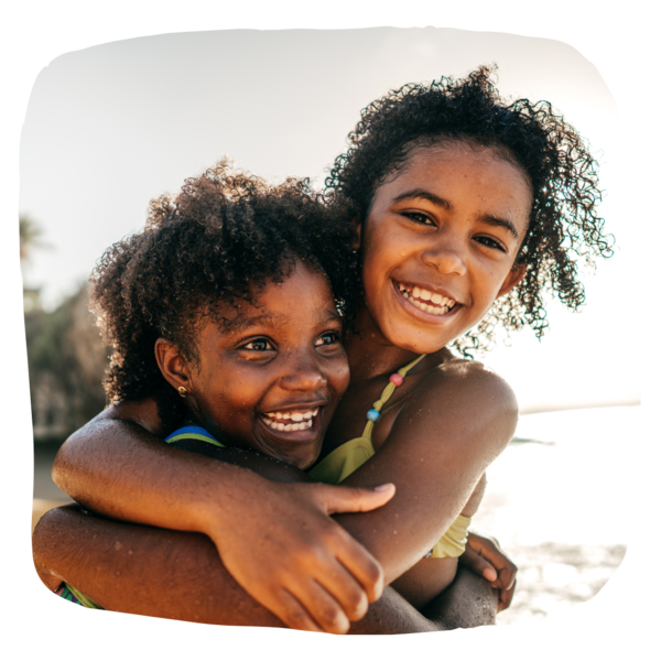 Two children on the beach hugging each other and smiling wide. Their coily hair is blowing in the wind. 