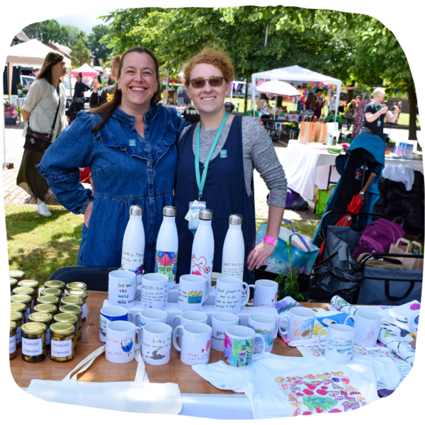 Two women smiling wide behind their market stall table full of tote bags, bottles and jars.