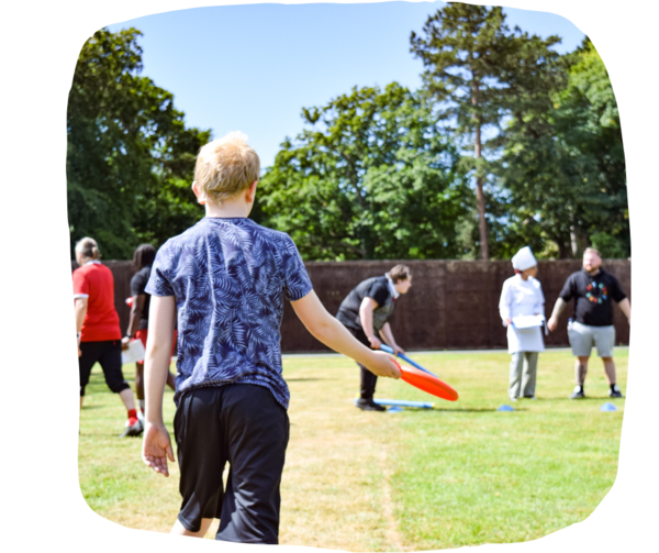 Young boy holding a frisbee walking away to throw it. in the background there are other teachers and students playing sports