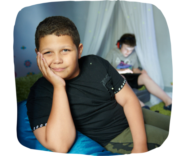 two boys sat down on beanbags in a room with low light. One is wearing noise cancelling earphones and playing on a tablet, while the other is smiling at the camera. There is sensory furniture around them including light up curtains and wall stickers.