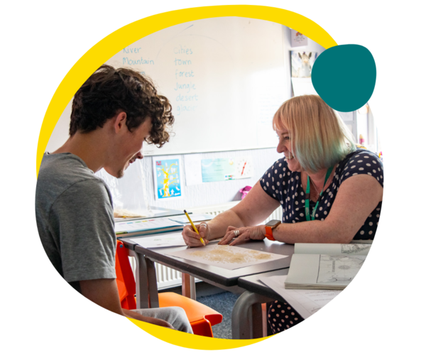A teen boy is laughing as he is completing coursework next to a teacher smiling at him. 