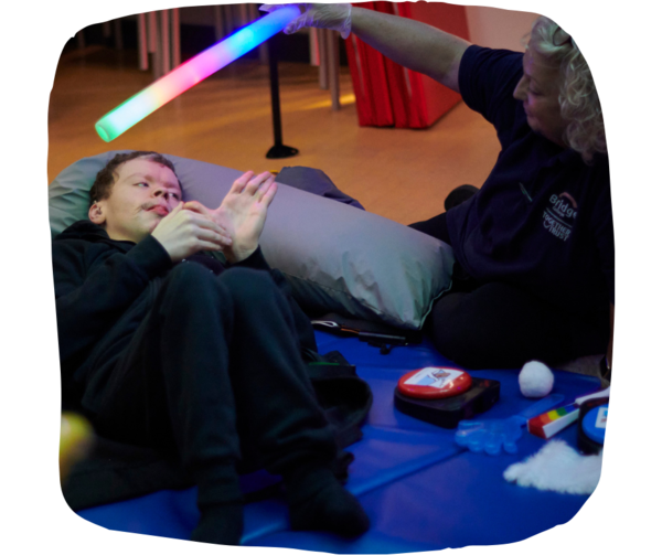 Man lying down on a soft mat on the floor in the sensory room. Woman next to him is holding up a light stick.