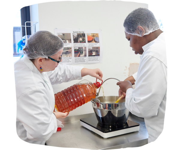 Man and woman wearing chef aprons and nets cooking soup in a big saucepan. The man is stirring with a wooden spoon and the woman is pouring liquid from a big jug. 