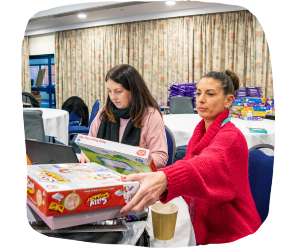 Two women wearing jumpers sitting at a table in a room full of toys and chocolate boxes.