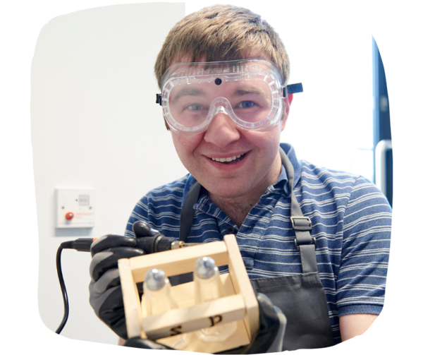 Student wearing protective goggles, holding a pyrography machine and a piece of wood, smiling at the camera.