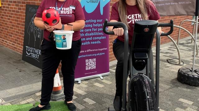 A team member from Booths holding a collection bucket next to another sat on an exercise bike
