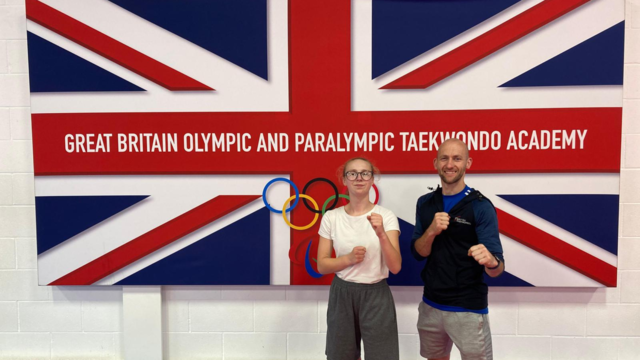 Teenage girl stood next to a man both holding up their fists in a fighting stance. They are stood in front of a Union Jack flag that reads "Great Britain olympic and Paralympic taekwando academy" 