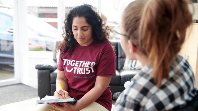 An adult woman sitting and speaking to a young lady whilst taking notes in a notebook.