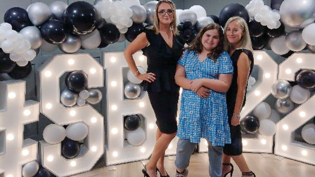 Alice wearing a blue dress standing in front of a light up sign alongside her 2 teachers wearing black dresses. They are all smiling wide. 