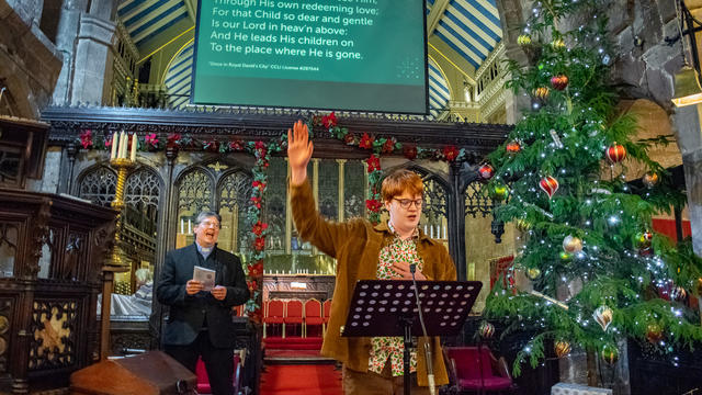 Teenage boy wearing a corduroy blazer singing. He is holding one hand up. He is stood in front of a music stand in an old church. Behind him is a screen with lyrics to the song. 