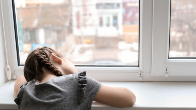 Young girl with her head resting on her arms, looking forlorn out of the window.