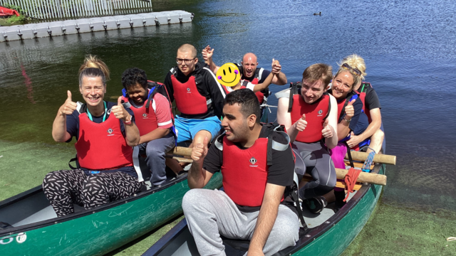 Two kayaks on the edge of the water full of students and staff wearing red life jackets, smiling and waving at the camera