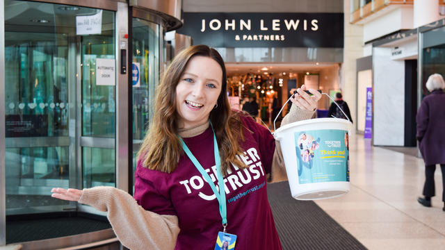 Woman wearing a purple together trust tshirt standing inside a John lewis store. She is holding a donation bucket and smiling wide. 