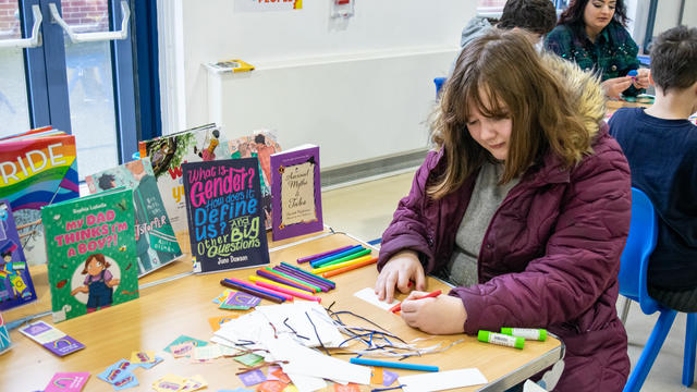 Young girl drawing on a bookmark at a table full of books and stickers. 