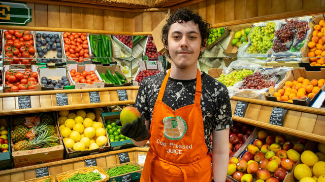 A teen boy wearing an apron is smiling in front of a well-lit wall of fresh fruit. He is holding a mango and smiling. 