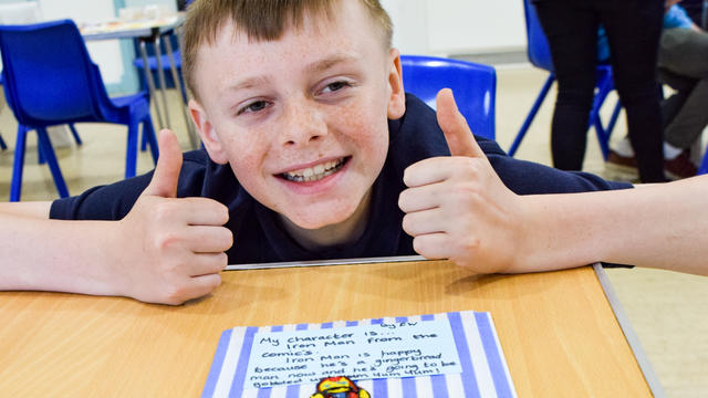 young boy smiling wide with both of his thumbs up. he is standing above a gingerbread man decorated to look like Iron Man