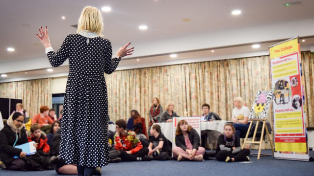 Woman wearing long dress standing in front of a group of children. She is talking to them pointing to a screen. The children are all sat on the floor looking up. 