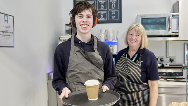 A student holding a tray with a drink on it with a member of staff stood behind