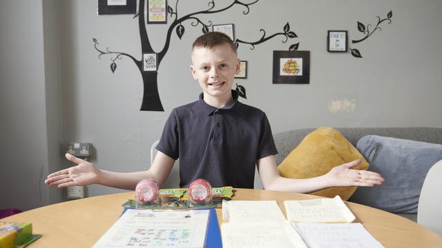 A student sat in front of some of their work smiling with their arms outstreched 