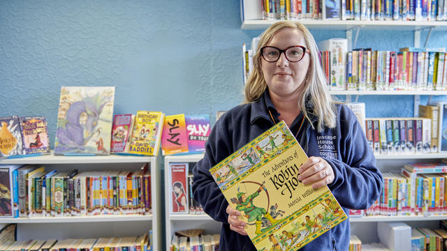 A member of staff holding a large yellow book smiling at the camera stood in front of a bookshelf