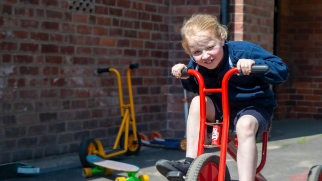 Young girl cycling on a red bike in a courtyard. She is winking at the camera as sun shines in her face