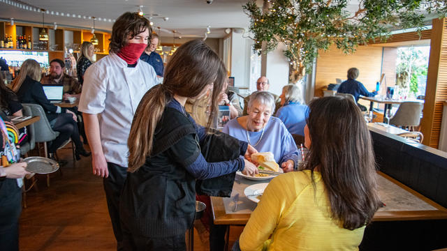 Two young people presenting a portion of pasta to a table of customers. One of them is grating cheese over the pasta. 