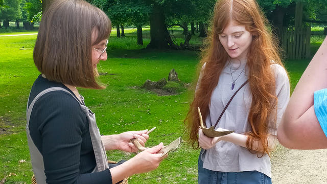 Young girl holding deer antlers. She is looking down smiling. Next to her is a staff member from Dunham Massey park explaining. They are stood outside in the park.