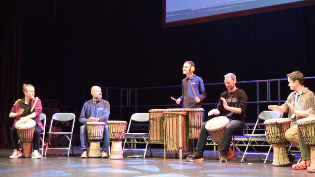 Students and staff in a row on stage. They are playing the drums.