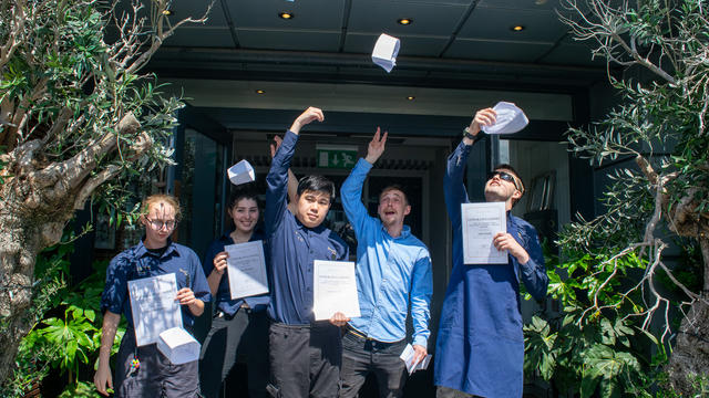 A group of 4 students and an employee from Gusto throwing up paper hats in the air outside the restaurant. 