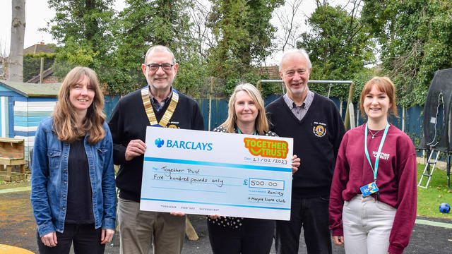 A group of 5 people smiling holding a cheque. They are stood outside in a garden. 