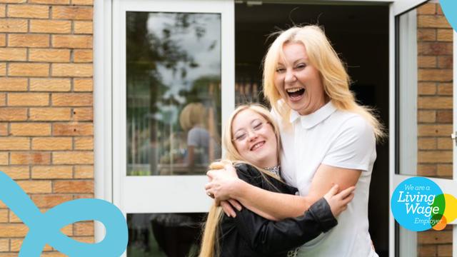 Two people smile at the camera, hugging each other. They are outside with a building behind them.