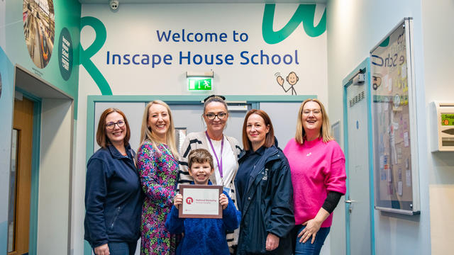 The Nurture team and a pupil, holding the Award certificate and posing for a photo in front of the "Welcome to Inscape House School" wall