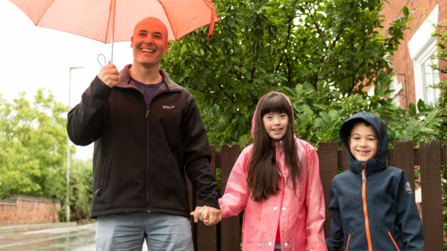 a man with two children smiling at the camera. They are holding hands and the man is holding an umbrella.