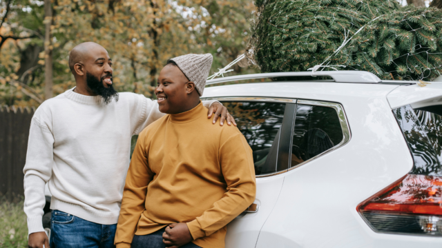 A man standing next to a teenager with one arm around his shoulder. The are outside leaning on a car. on top of the car is a christmas tree. 