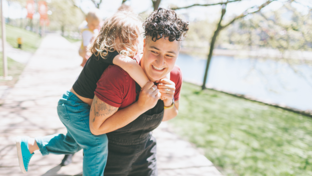 Woman holding a child on her back as they're both laughing in a park. 