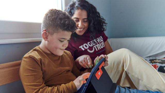 Young boy with hearing aid sitting down on his bed holding a tablet. An adult woman is sat down next to him smiling as she is helping him with the tablet. 