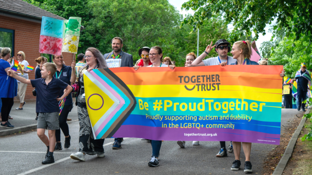 a Group of teachers walking outside a school in a march holding a rainbow banner that reads "be #proud together. proudly supporting autism and disability in the LGBTQ+ community" 