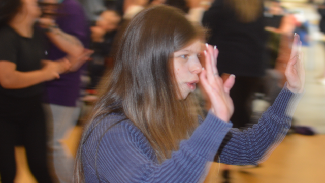 A young woman dancing in a crowed hall. Her hands are in the air. 