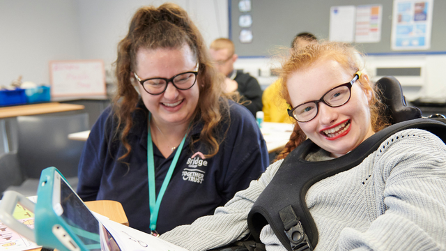 Student in wheelchair and teacher sitting down in class smiling. The student is holding a tablet. 