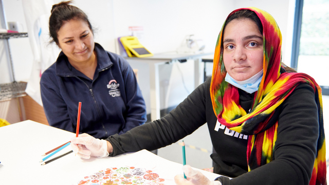 Student wearing a colorful head scarf drawing. Next to her is a teacher smiling at her.